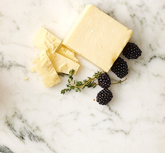 Block and slices of Murray’s High Plains Cheddar cheese, blackberries, and a sprig of thyme on a white marble surface.