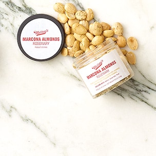 Canister of Murray’s Marcona Almonds with Rosemary spilling out onto a white marble surface.