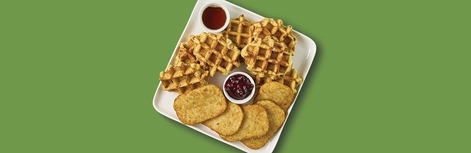 White platter with hashbrowns and waffles and dipping bowls of maple syrup and cranberry sauce. Platter is on a green back-ground.