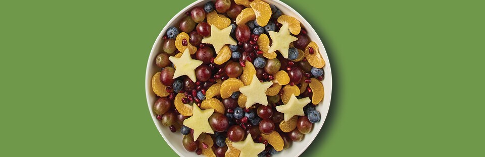 Fruit salad in white bowl. Salad has grapes, blueberries, orange segments, pome-granate arils and apples cut into star shapes. Salad is on a green background.