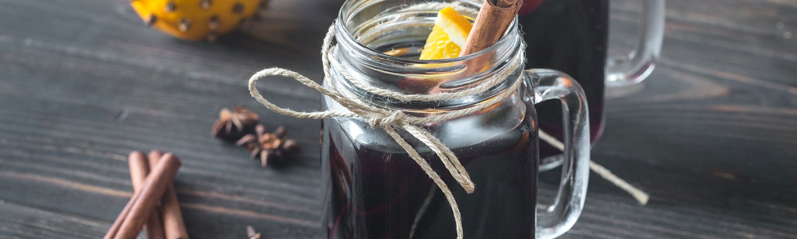 On a rustic wooden table two mason jars sit, wrapped in twine and filled with a dark beverage, an orange slice, and a cinnamon stick
