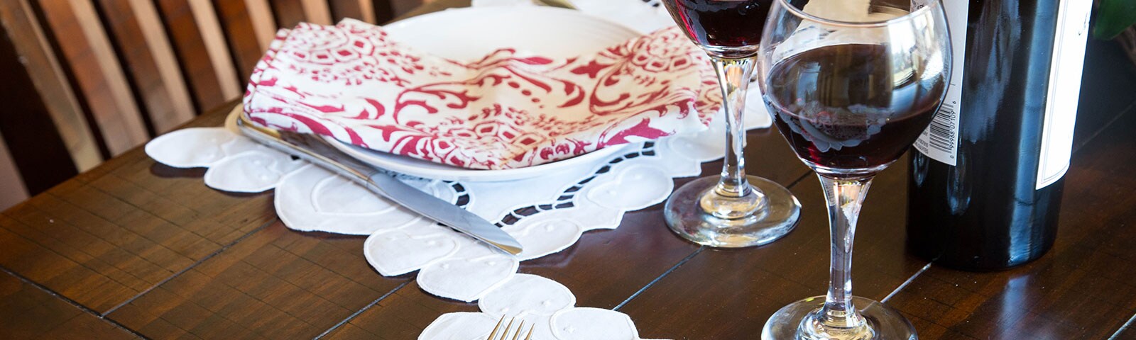 A wooden table, atop sits two wine glasses filled with red wine and two place settings A wooden table, atop sits two wine glasses filled with red wine and two place settings