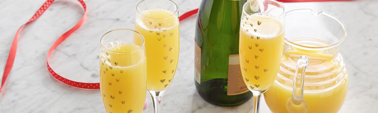 Crafty champagne flutes filled with mimosas in the foreground and candy hearts scattered on a marble countertop 