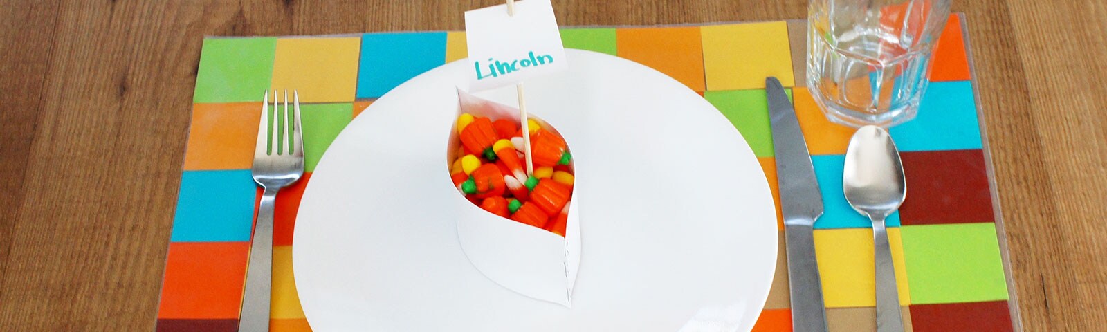 A colorful grid placemat on a wooden table with a white plate and a small jar filled with candy corn on top A colorful grid placemat on a wooden table with a white plate and a small jar filled with candy corn on top