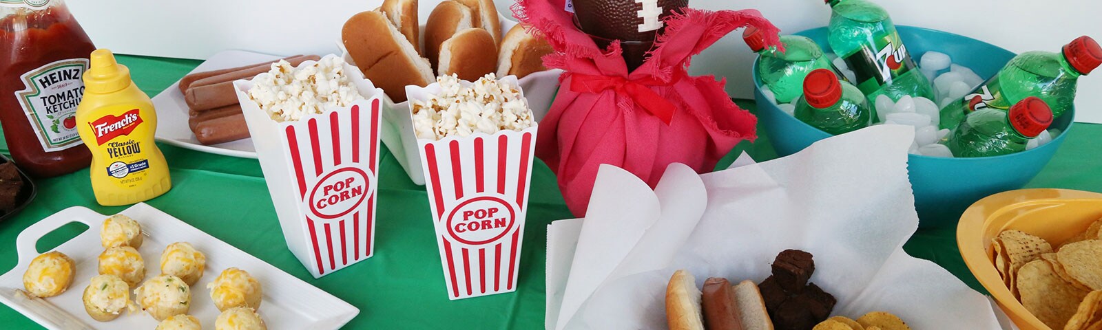 An array of football ready treats including popcorn, hotdogs and chips on a table covered with a green tablecloth An array of football ready treats including popcorn, hotdogs and chips on a table covered with a green tablecloth