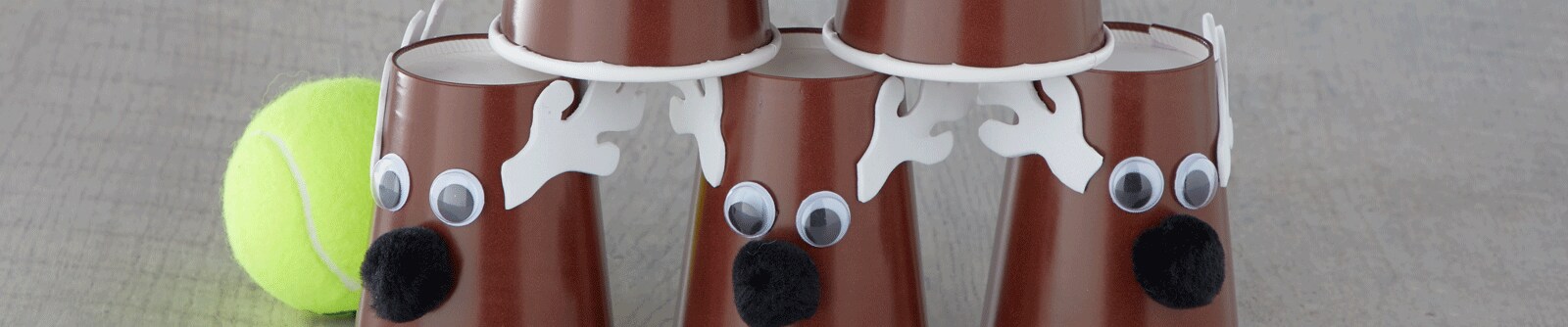 A stack of DIY reindeer using brown plastic cups with googly eyes, felt antlers and pompoms next to a tennis ball