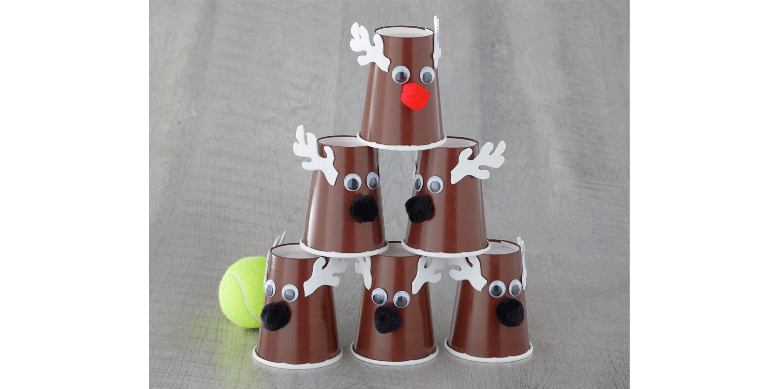 A stack of DIY reindeer using brown plastic cups with googly eyes, felt antlers and pompoms next to a tennis ball